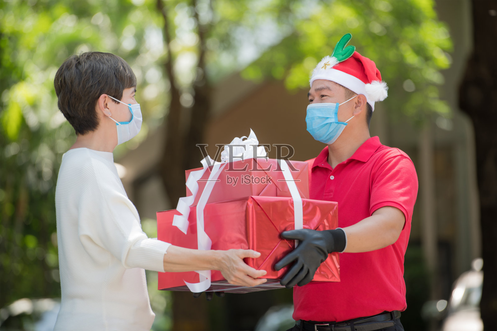 2019冠状病毒肺炎疫情期间，一名身穿红色制服、戴着口罩和手套、戴着圣诞帽的亚洲快递员在圣诞节期间为女性收件人递送礼物和礼品盒照片摄影图片
