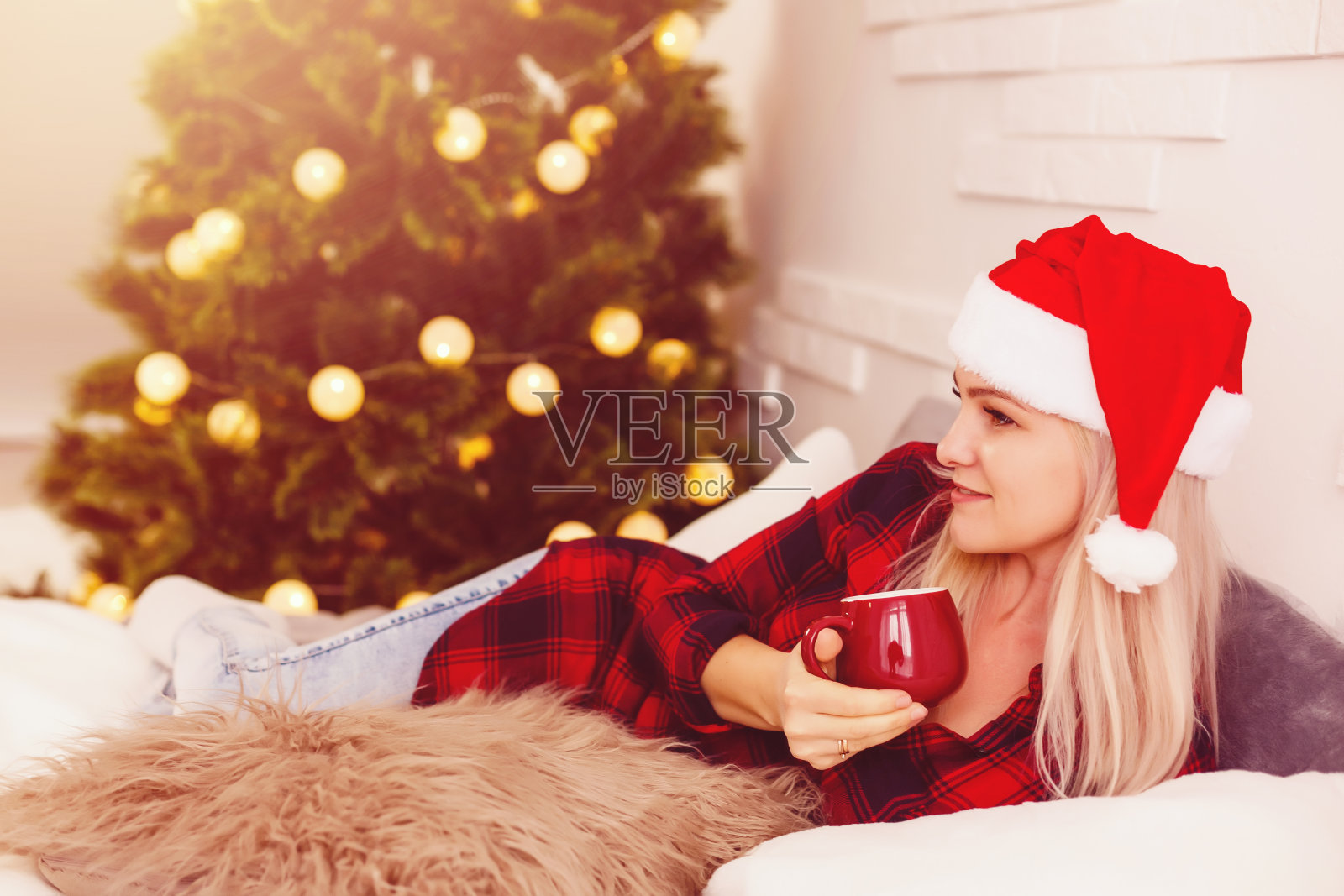 美丽微笑的女孩穿着温暖的新年毛衣坐在温暖的白色地毯上，手里拿着一杯热茶，旁边是装饰好的圣诞树和礼品盒。照片摄影图片