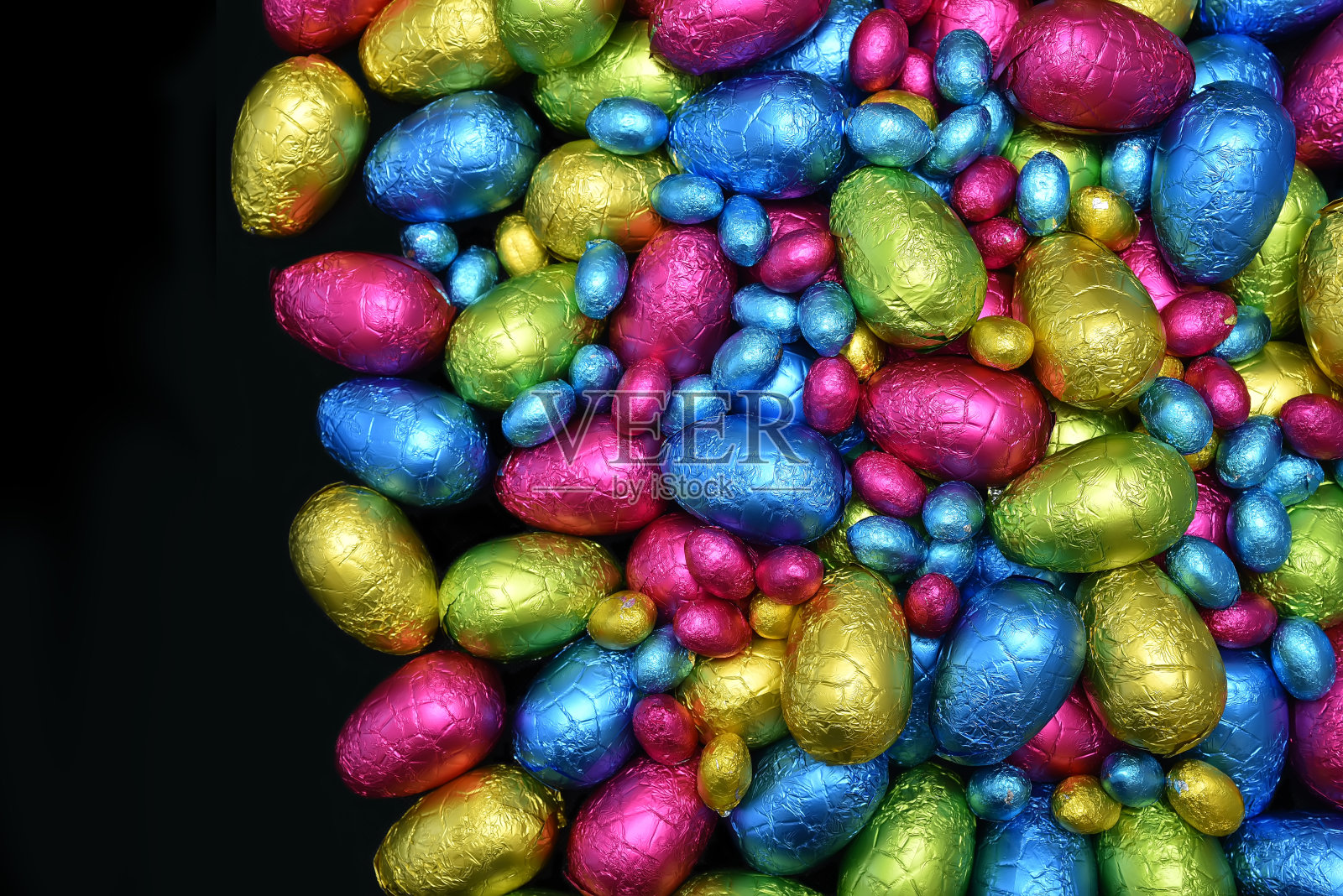 用粉红色、蓝色、黄色和酸橙绿色的彩色锡箔纸包成一堆或一组巧克力复活节彩蛋，背景为黑色，并留有空白。照片摄影图片