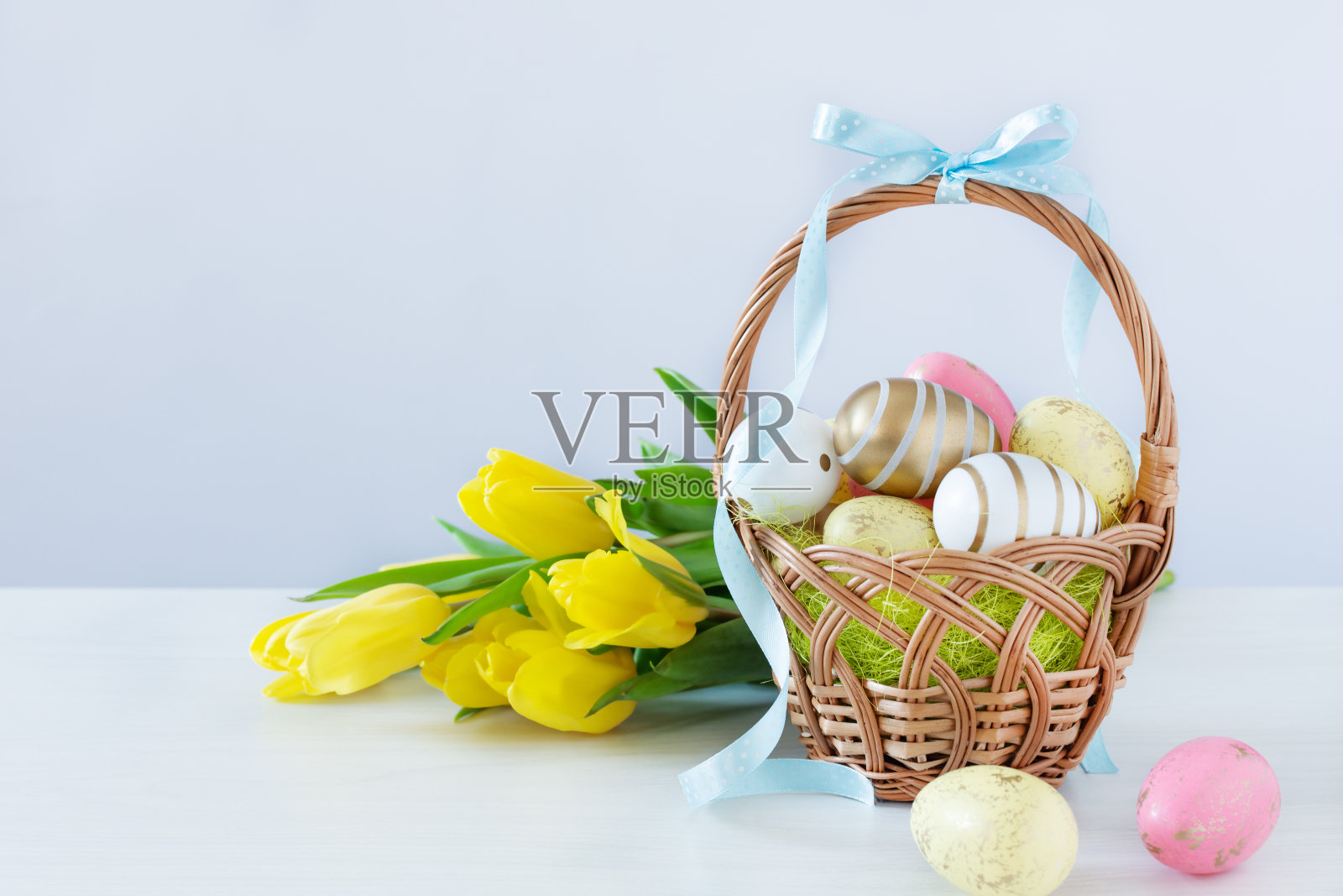 白色的木桌上放着一篮彩色的复活节彩蛋，上面放着郁金香花，背景是白色。照片摄影图片