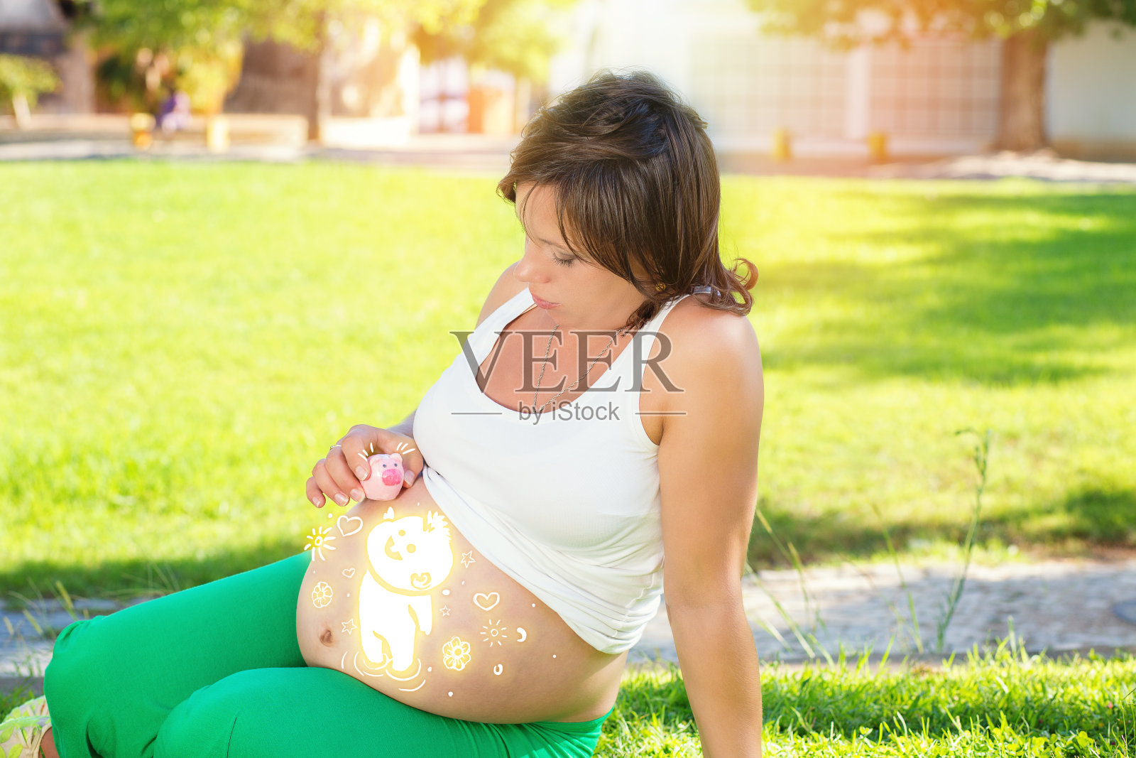 一个怀孕的妈妈正抱着存钱罐坐在公园里的绿草地上照片摄影图片