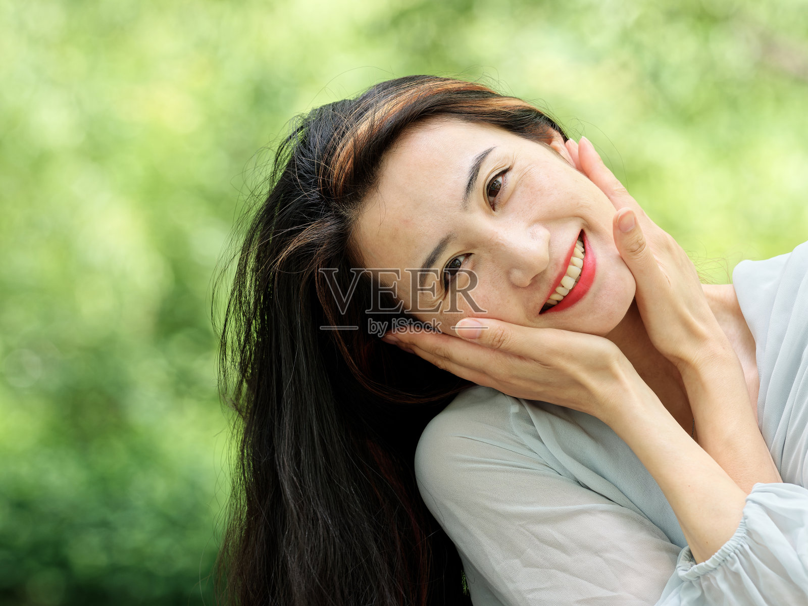 美丽的中国女人微笑在相机与她的手在她的脸，近距离肖像女性模糊的自然背景。照片摄影图片