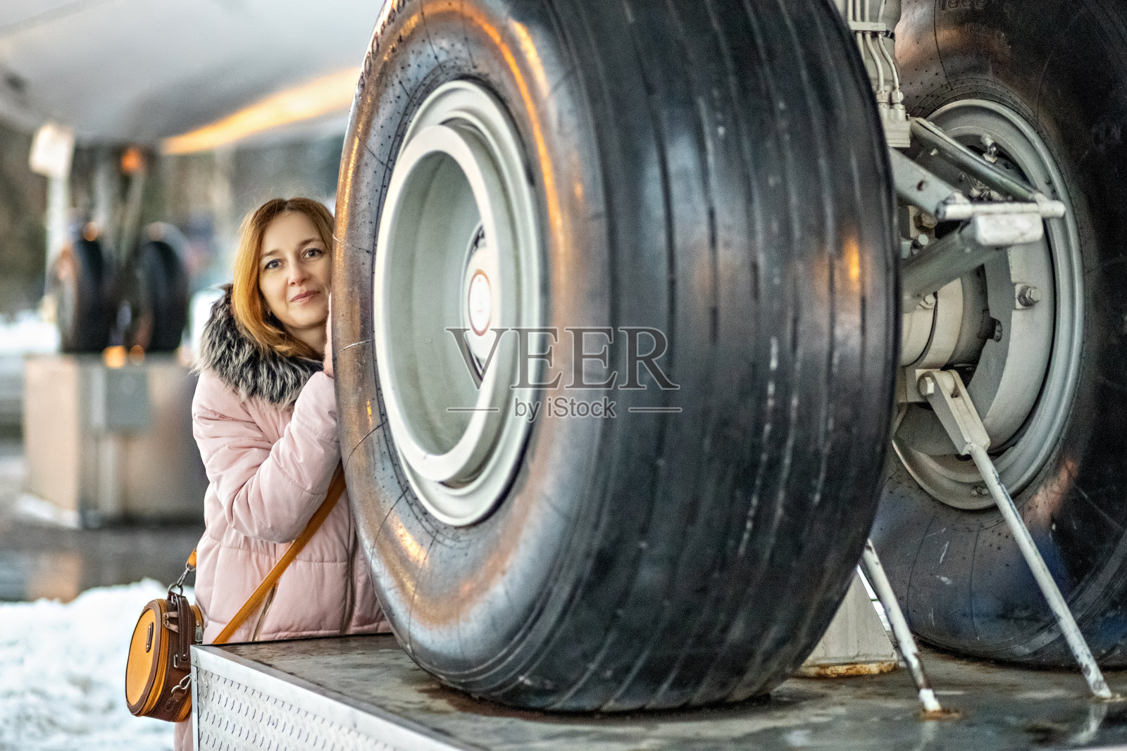 一个年轻的女人从大车轮后面向外张望。在机场修理的一架客机的前起落架照片摄影图片