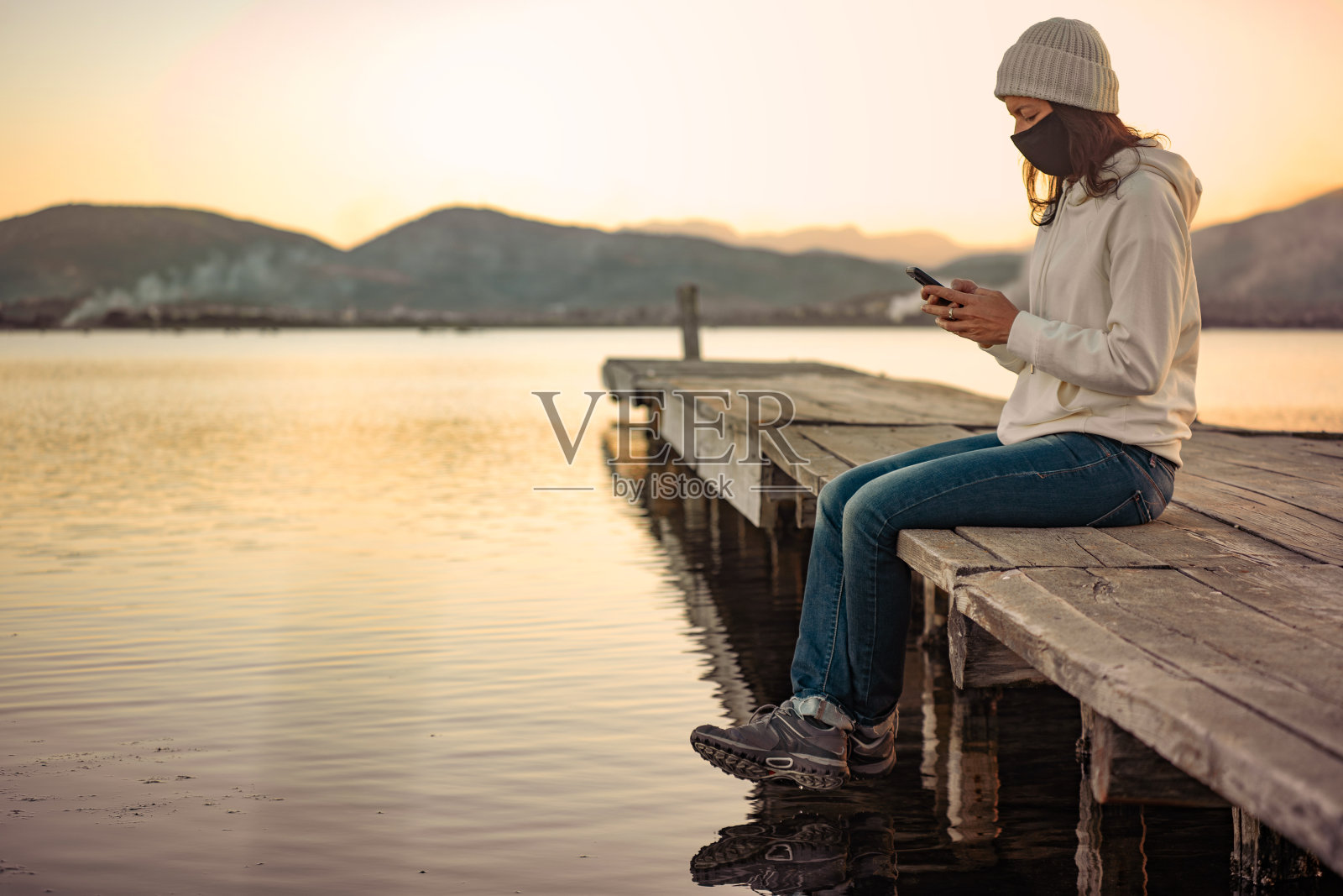 孤独女子在日落或日出时戴着防护口罩坐在码头上，用智能手机关注新冠肺炎的最新消息。互联网移动技术与冠状病毒新常态照片摄影图片