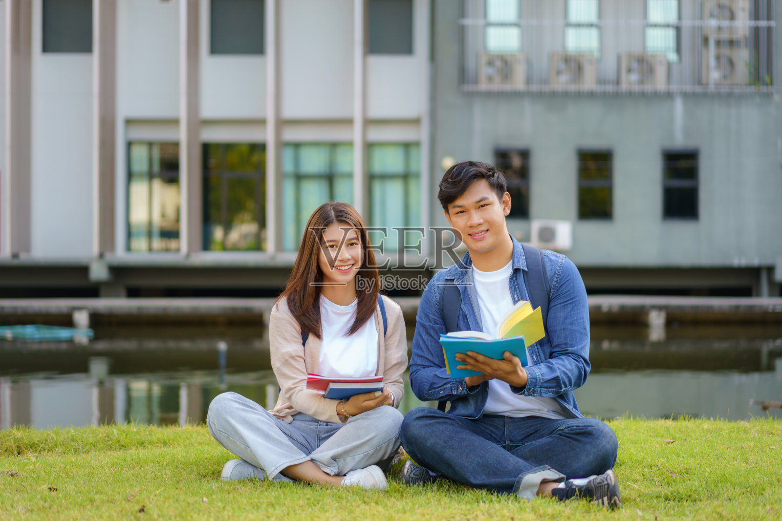 亚裔男女大学生坐在校园草地上看着镜头，微笑着，在公园里一起看书。照片摄影图片