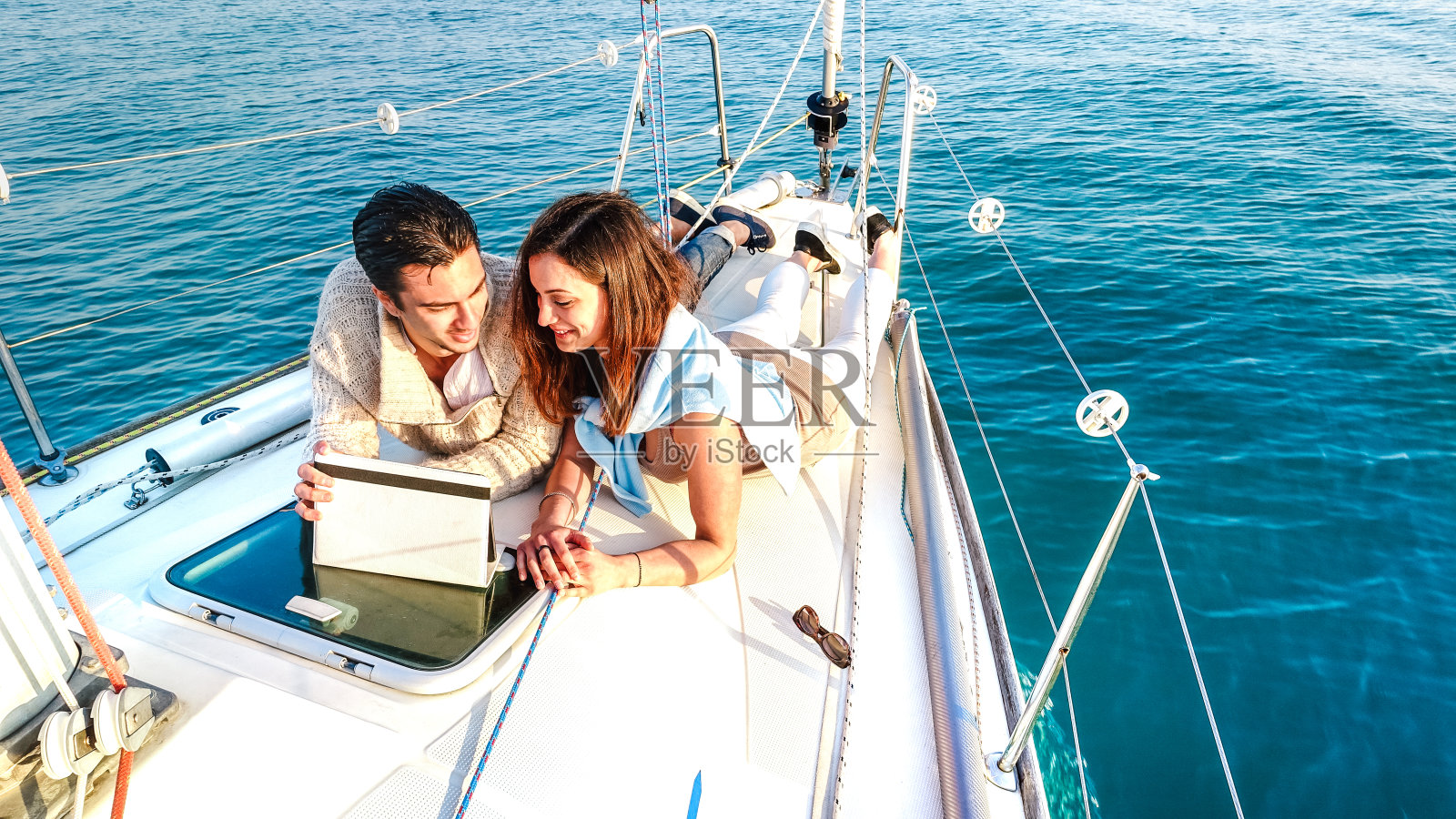 相爱的年轻夫妇在帆船上享受平板电脑的乐趣-豪华旅行生活方式和数字游牧概念的专属游艇旅游-明亮生动的滤镜与软鱼眼镜头失真照片摄影图片