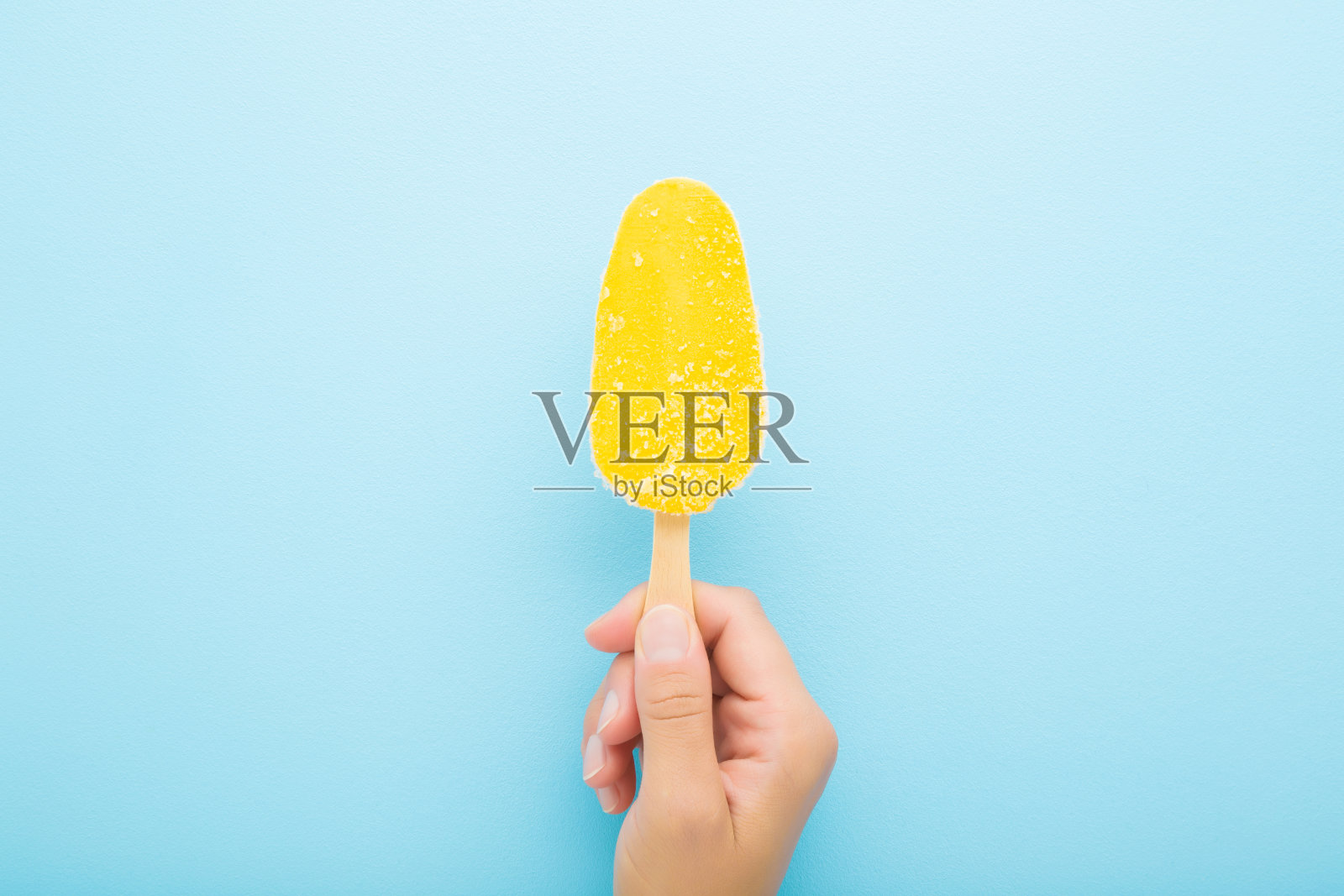 年轻的成年妇女手拿明亮的黄色芒果冰淇淋与棍子在浅蓝色的桌子背景。柔和的颜色。特写镜头。自顶向下的观点。照片摄影图片