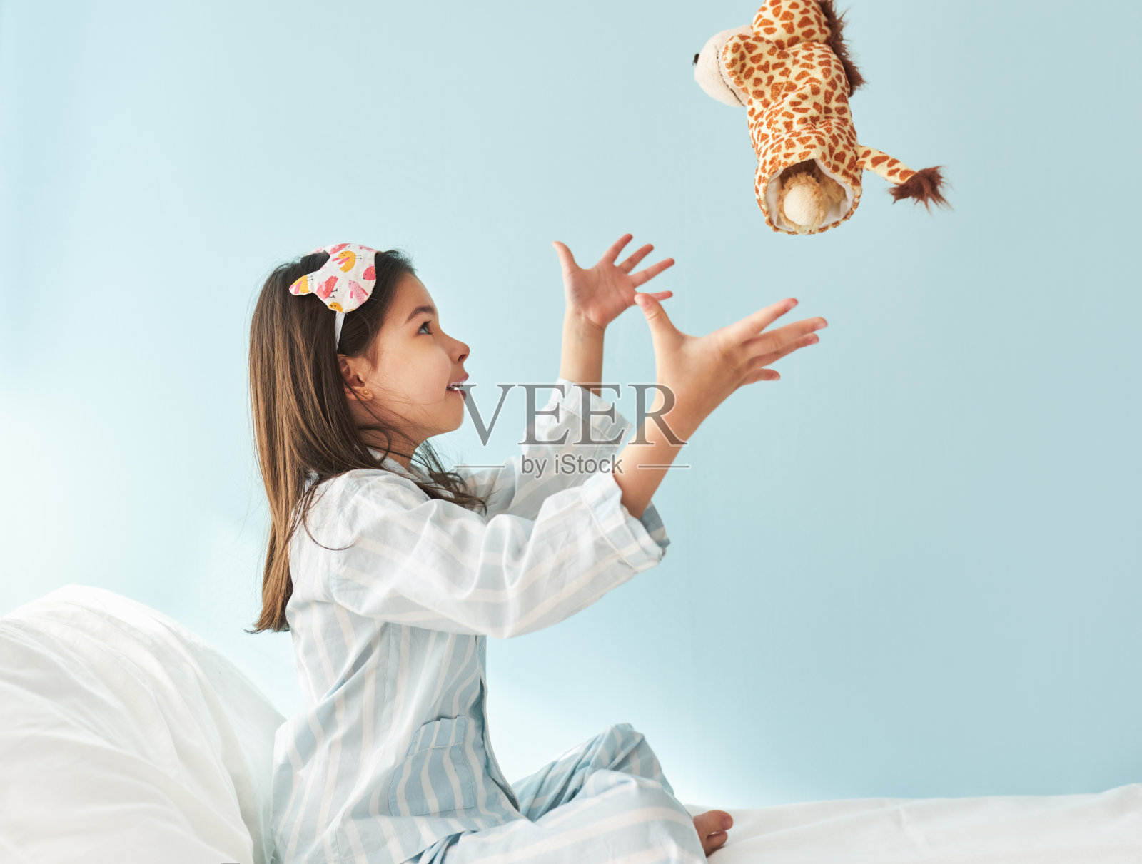 可爱的小女孩穿着蓝色的白色条纹睡衣，头戴睡觉面具，面带微笑，在床上玩玩具长颈鹿。孩子在早上有快乐的表情。照片摄影图片
