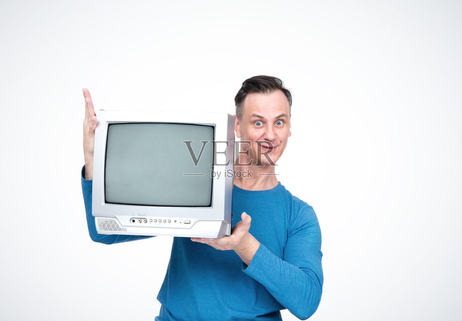 一个穿着蓝色t恤的快乐微笑的男人，手里拿着一台旧的CRT电视，在灯光背景上。照片摄影图片