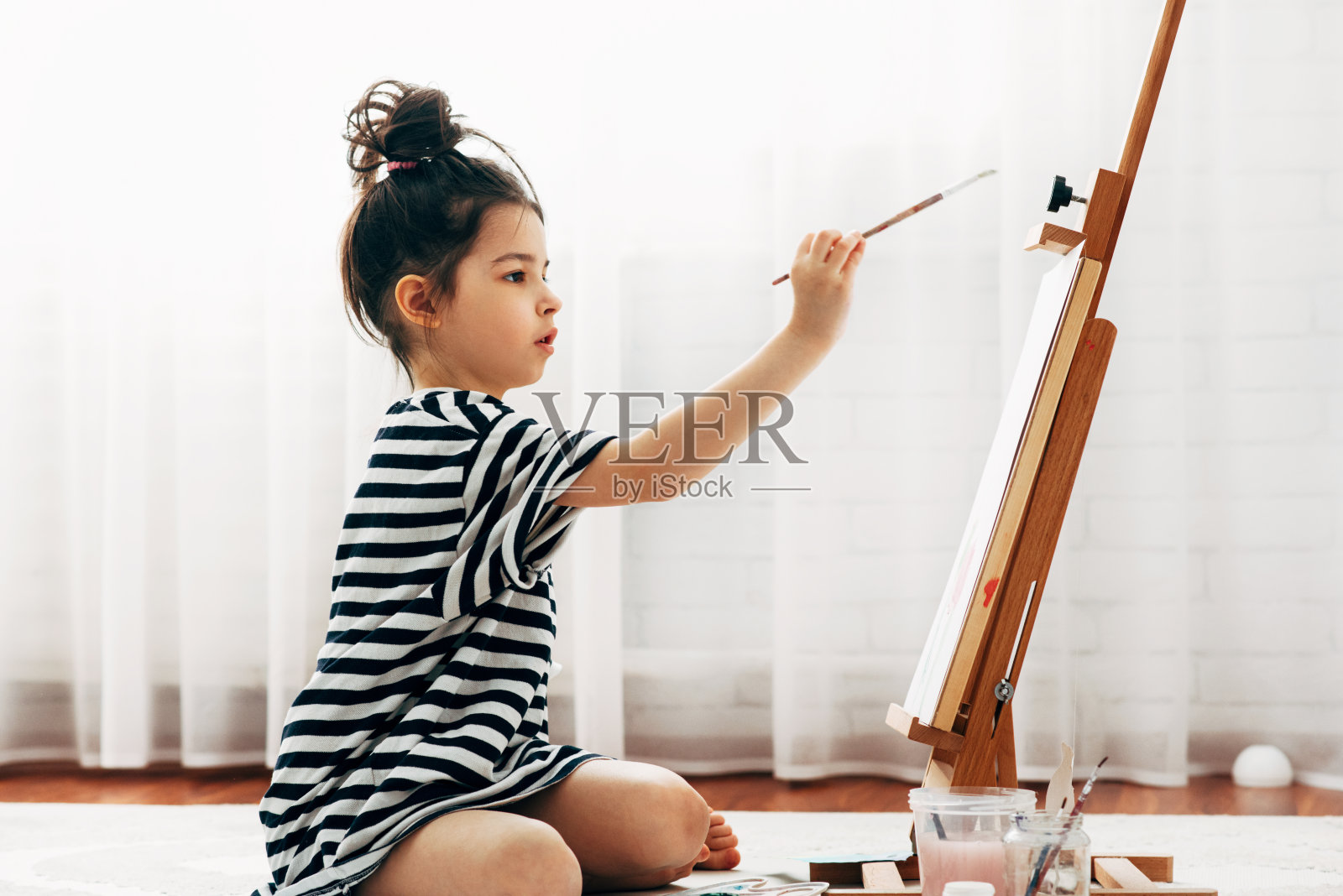 一个学龄前的小女孩在家里的画架上用画笔在纸上作画。一个孩子坐在地板上，在她的房间里画画，寻找想象力。照片摄影图片