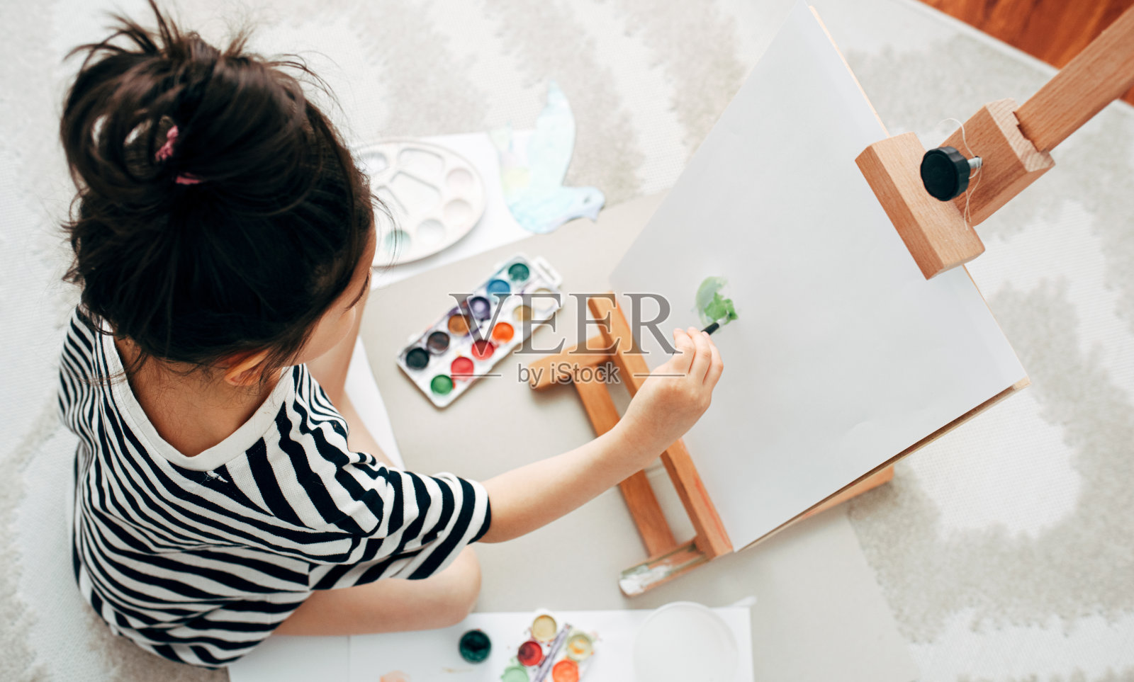 上图是一个小女孩在家里的画架上作画。女学生坐在地毯上，在她的房间里画画。照片摄影图片