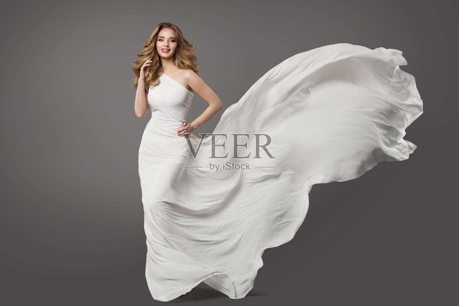 穿白裙子的女人。美丽的新娘模型在婚礼礼服上的风在灰色的背景。新娘时尚服装，卷曲的发型和化妆照片摄影图片