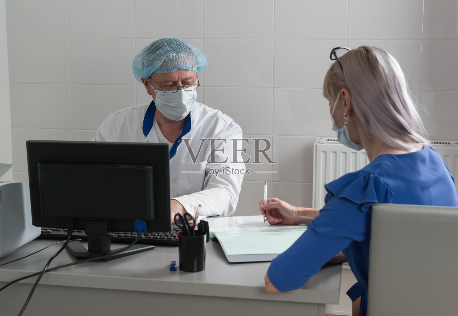 一名身穿白大褂、戴着口罩和帽子的医生坐在医疗办公室的桌旁，看着患者签署知情同意书。有选择性的重点照片摄影图片