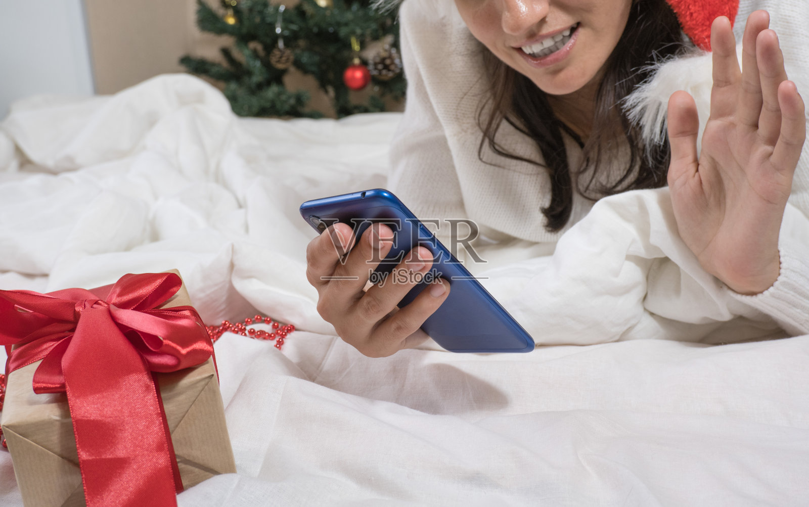 美丽快乐的黑发女人穿着白色毛衣，戴着圣诞帽，躺在白色的床上拿着圣诞礼物，用手机自拍照片摄影图片