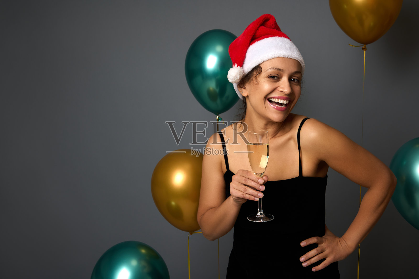 美丽的年轻女子戴着圣诞老人的帽子，身穿黑色晚礼服，手持香槟长笛，在灰色的背景和闪亮的金色和绿色的气球上庆祝圣诞节照片摄影图片