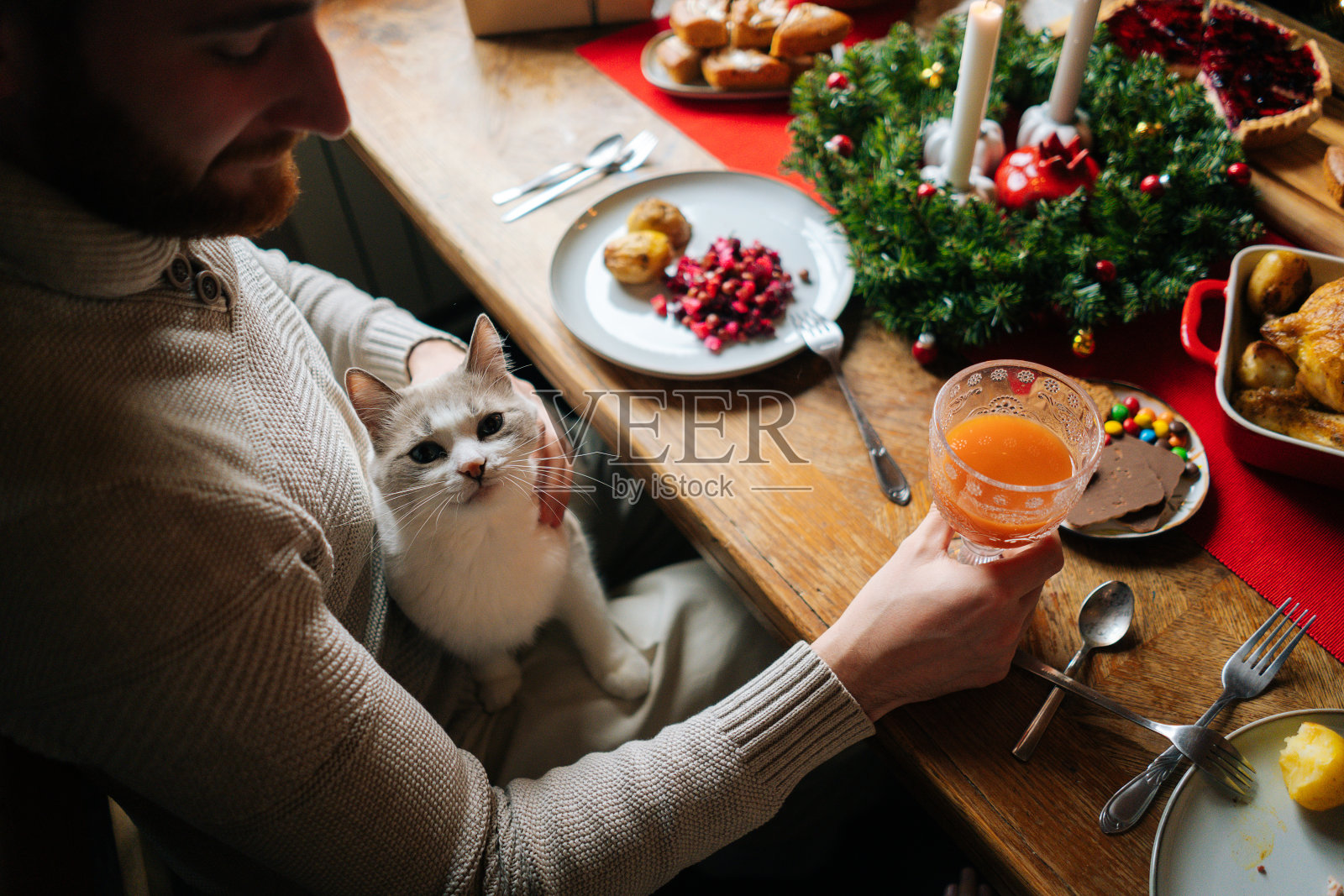 在节日家庭聚会期间，长着胡子的英俊年轻人坐在餐桌旁，腿上放着一只猫，手里拿着一杯新鲜果汁。照片摄影图片