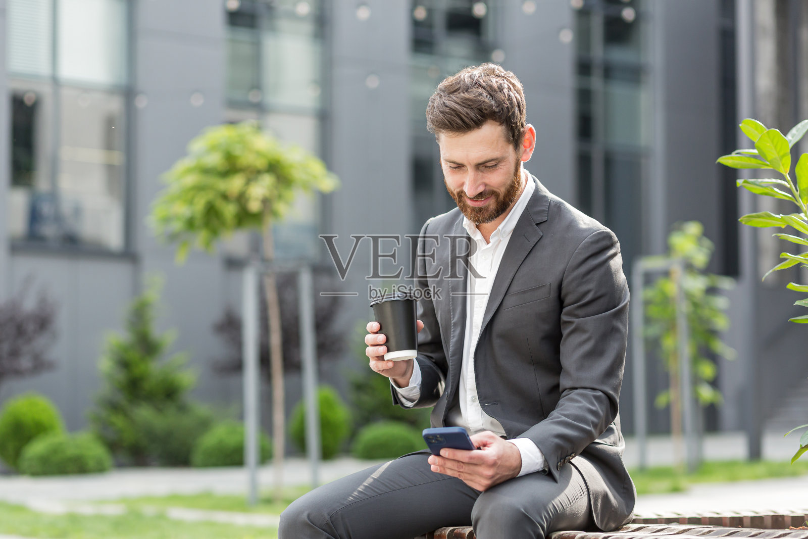 英俊快乐的大胡子商人坐在长凳上，端着杯咖啡，用手机浏览手机，读着喜讯，开心的笑着。外面有现代都市街道的背景。城市公园照片摄影图片