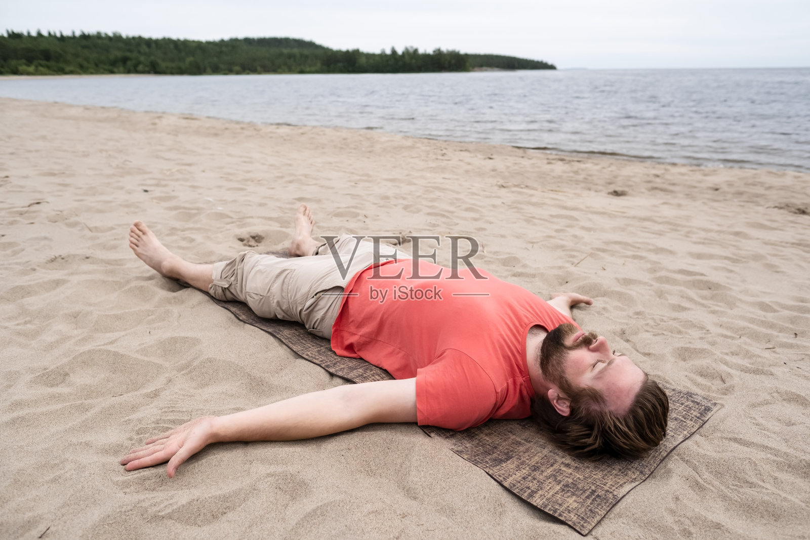 一个长胡子的男人在做瑜伽，他做Savasana练习，躺在湖边的沙滩上，以身体姿势放松。照片摄影图片