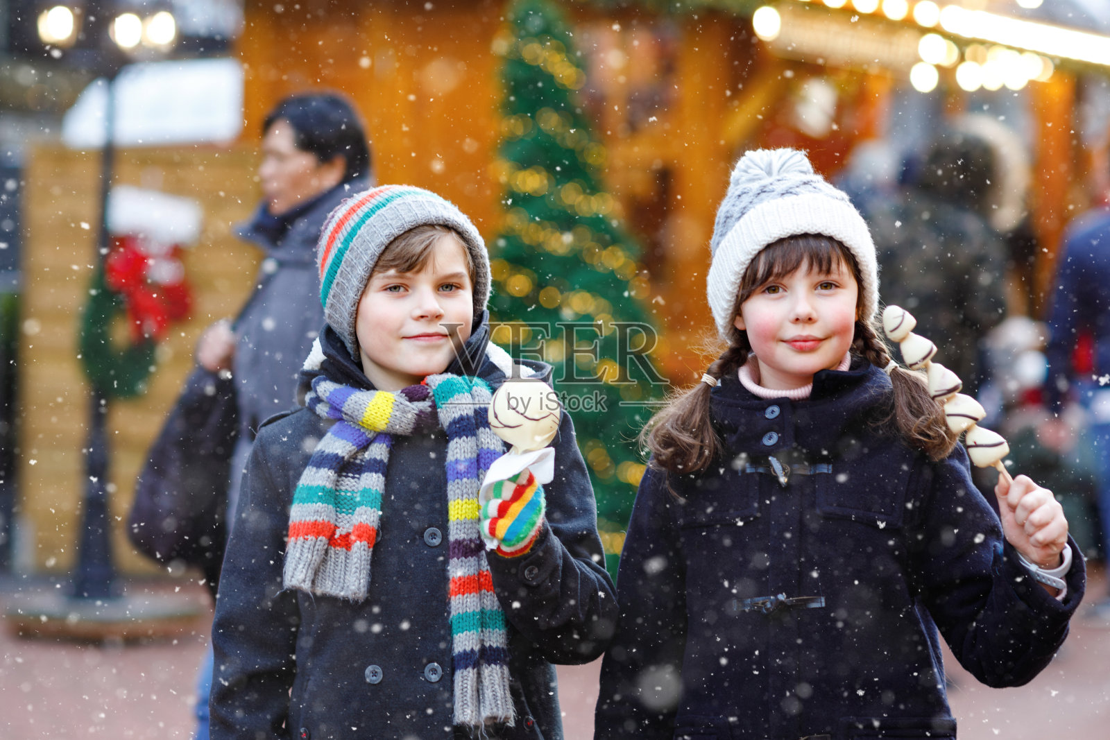 在传统的德国圣诞市场上，可爱的小女孩和小男孩在吃着串着草莓和苹果的白巧克力。快乐的孩子，最好的朋友，双胞胎和兄弟姐妹在下雪天照片摄影图片