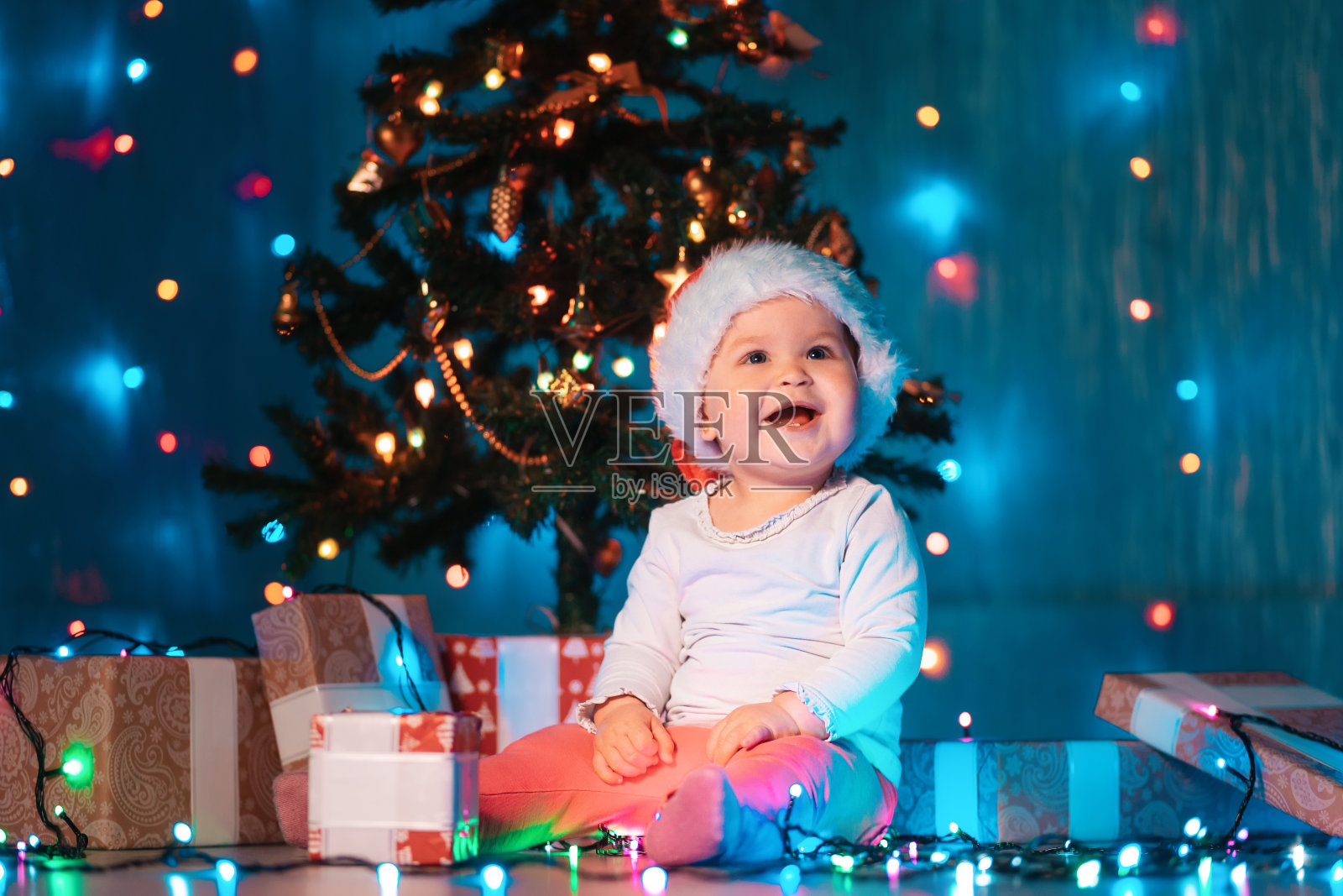 快乐的宝贝女孩戴着圣诞帽坐在节日的灯光和圣诞礼物。室内。冬季假期的概念照片摄影图片