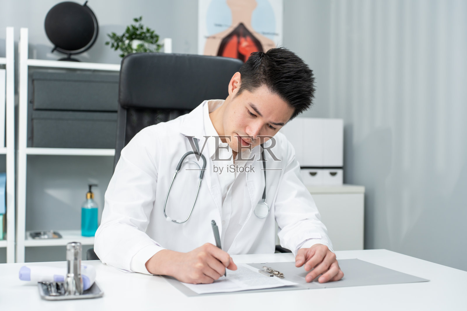 亚洲专科男医生在办公室医院的桌子上写纸条。有吸引力的医生使用笔记本电脑工作，诊断治疗病人的人在虚拟在线远程保健。照片摄影图片