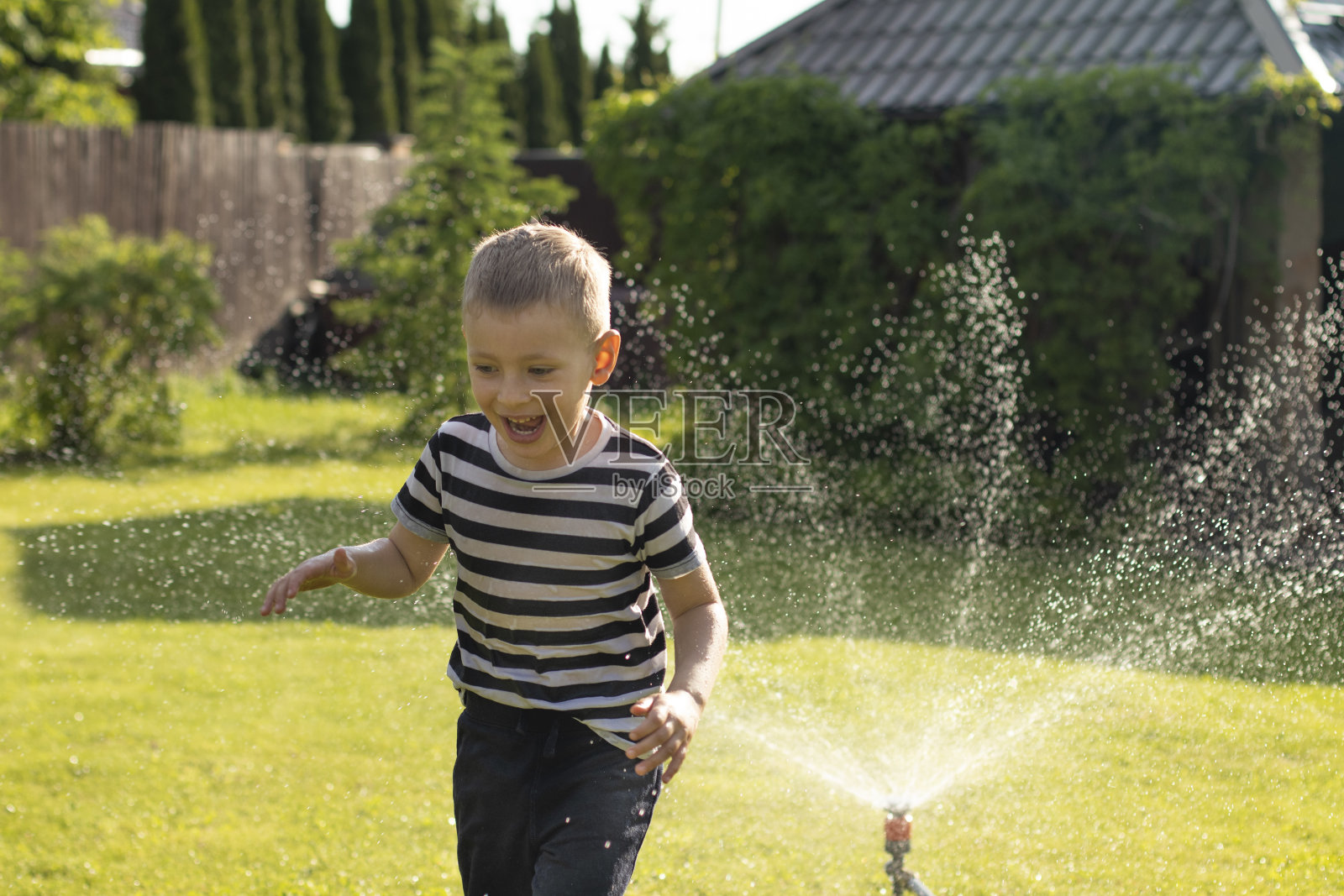 一个浑身湿透的小男孩光着脚在洒水器旁边的草坪上奔跑。快乐无忧无虑的童年和假期概念。照片摄影图片_ID:423126989-Veer图库