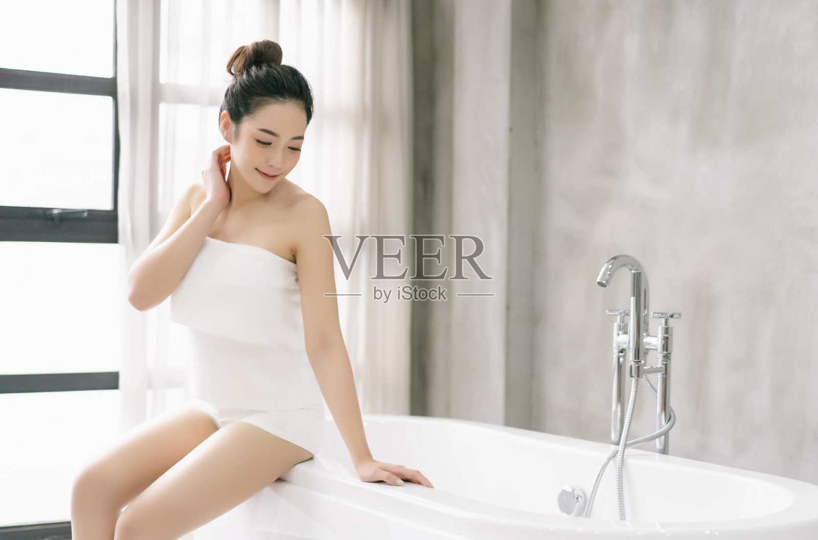 美丽的微笑的亚洲妇女是放松坐在一旁的独立的白色浴缸在浴室。最喜欢的沐浴仪式，治愈浴，幸福，卫生和放松的概念。照片摄影图片
